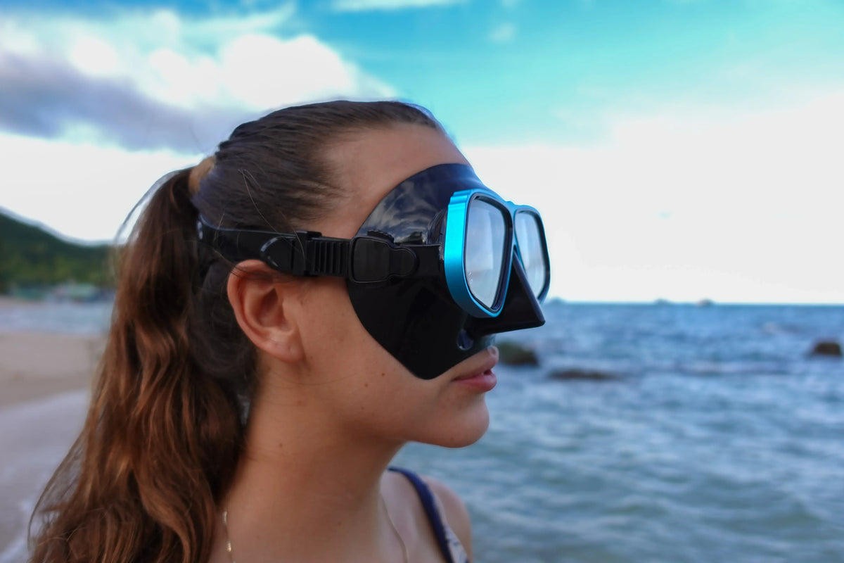 Side view of woman wearing Prescription scuba dive mask with Aluminium Blue frame and Black seal. The Rx Lumix mask. Standing in front of the ocean. 